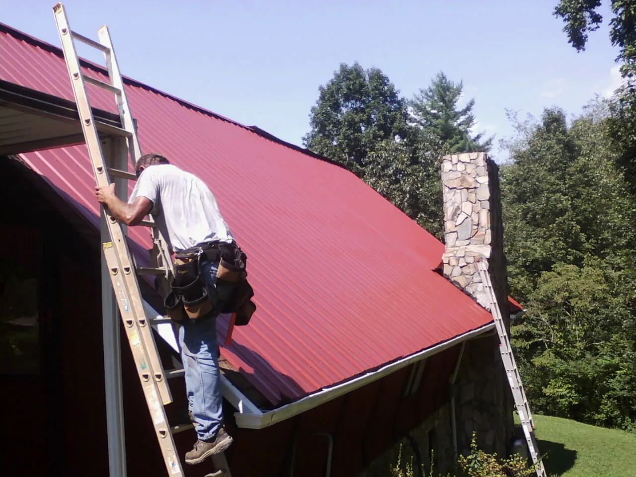 Roofer on a ladder beside a red metal roof and stone chimney — Soffit & Fascia Repair by Roofing Brothers in Union Mills